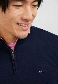 Pull à col zippé marine en tissu tricoté fin avec un col intérieur rose, orné d'un petit logo sur la poitrine et présentant une texture lisse.