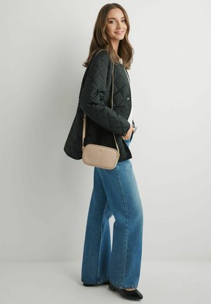 Młoda kobieta uśmiecha się, ubrana w czarną pikowaną kurtkę, niebieskie spodnie o szerokich nogawkach, czarne buty i nosząca beżową torebkę na ramię.