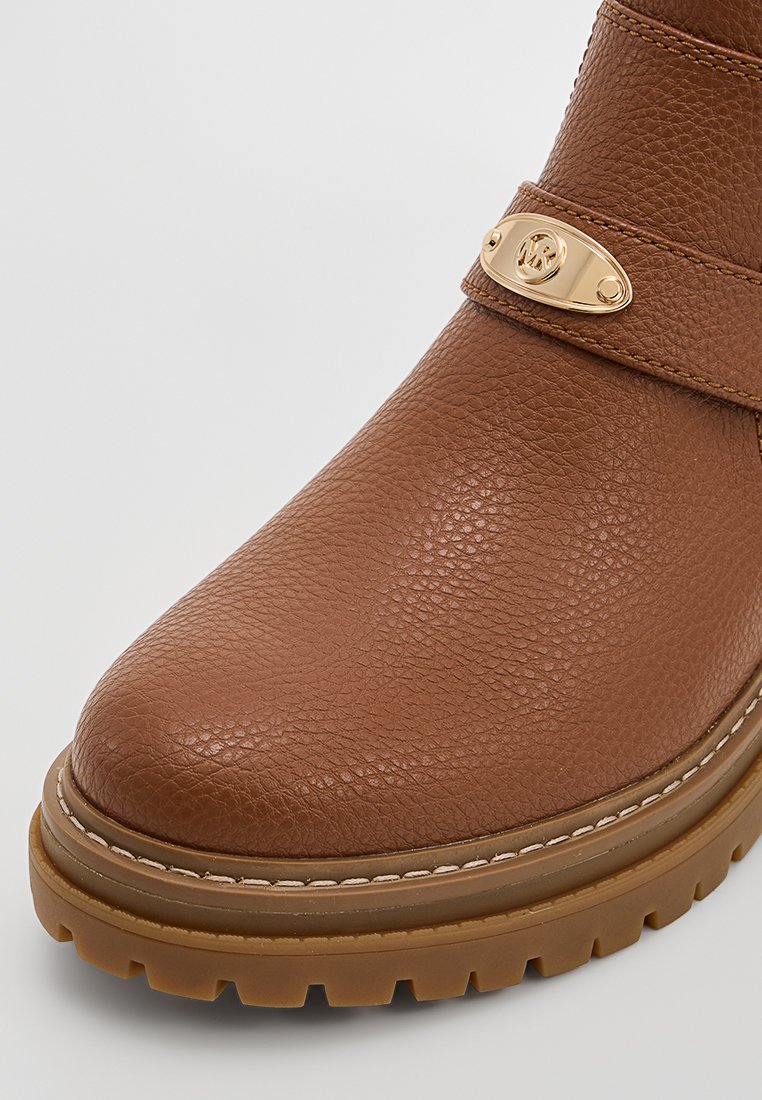 Bruine gestructureerde leren laars met een dikke rubberen zool en gouden ovale metalen detail op de strap over de voorkant.