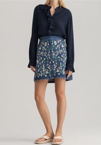 Blusa azul marino con cuello acampanado, combinada con falda de mezclilla azul con bordado floral y corte recto. Chanclas neutras completan el look.