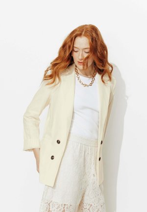 Femme aux cheveux roux portant un blazer crème, un haut blanc et un pantalon en dentelle, regardant vers le bas avec les cheveux flottant doucement d'un côté.