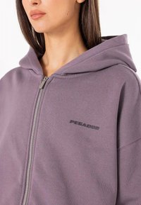 Fialová mikina na zips z mäkkej látky, s kapucňou na šnúrky a malým čiernym logom na hrudi. Hladká štruktúra a pohodlný strih.