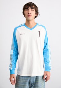 Jersey à manches longues blanc avec des accents bleu clair, présentant un col en V, un grand numéro "1" et trois rayures blanches sur les manches.