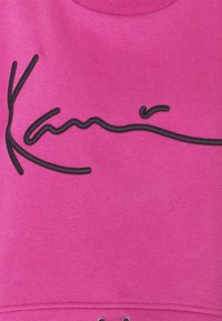 Sudadera fucsia con cuello redondo, presenta un diseño de firma bordado en negro y una tela texturizada. Incluye detalles de ojales metálicos.