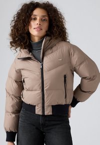 Chaqueta tipo abrigo en marrón claro con cierre de cremallera, bolsillos laterales y puños acanalados. Presenta un diseño acolchado y un logo en el lado izquierdo del pecho.