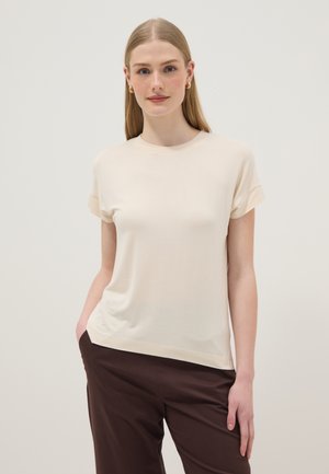 Vrouw met lang blond haar die gouden creolen draagt, een crème top met korte mouwen en donkerbruine broek, staand met één hand in de zak.