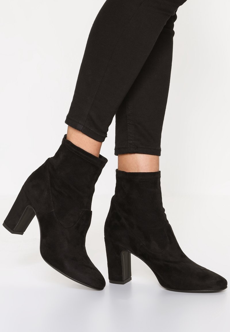 Personne portant des bottines en daim noir à talons carrés et un pantalon noir ajusté sur fond blanc.