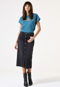 T-shirt turquoise avec le motif "AMORE", associé à une jupe en denim noire mi-longue avec des boutons avant et une fente sur le côté, portée avec des bottines noires.