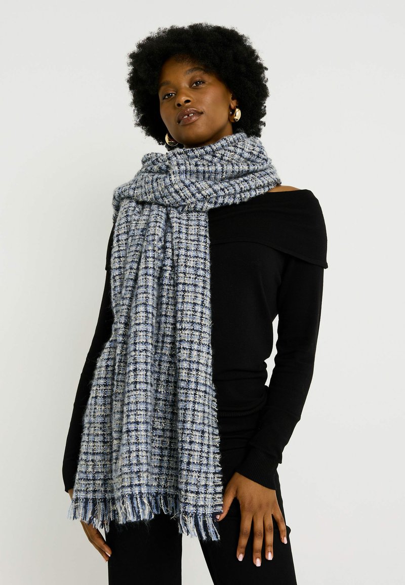 Bufanda tejida en azul y gris con un patrón de cuadros, bordes con flecos y una textura suave, llevada sobre una blusa negra sin hombros.