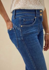 Mėlynos džinsinės kelnės su aukštu liemeniu, papuoštos auksinės spalvos mygtukais, šoninėmis kišenėmis ir tekstūruota medžiaga. Siuvinėtos detalės išilgai siūlių.