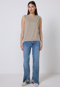 Top sin mangas color beige con texto bordado, combinado con jeans acampanados de color azul claro que tienen aberturas laterales y zapatillas blancas con detalles en negro.