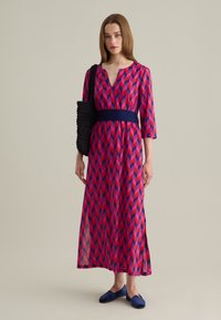 Maxikleid mit V-Ausschnitt, das ein geometrisches Muster in Pink, Rot und Dunkelblau zeigt. Ergänzt durch eine figurbetonte Taille und seitliche Schlitze. Wird mit navyblauen Schuhen getragen.