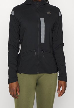 Osoba ubrana w czarną kurtkę z zamkiem błyskawicznym, z pionowym, odblaskowym paskiem i logo Adidas oraz oliwkowozielone spodnie, stojąca na białym tle.