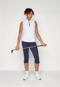 Camisa de golf sin mangas, blanca y con detalles en azul, combinada con pantalones capri en navy y zapatillas blancas. Sostiene un palo de golf con una mano.