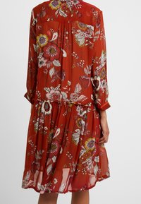 Robe florale rouge rouillée à manches longues et taille froncée, ornée de grandes fleurs multicolores dans des tons de rose, blanc, jaune et violet.