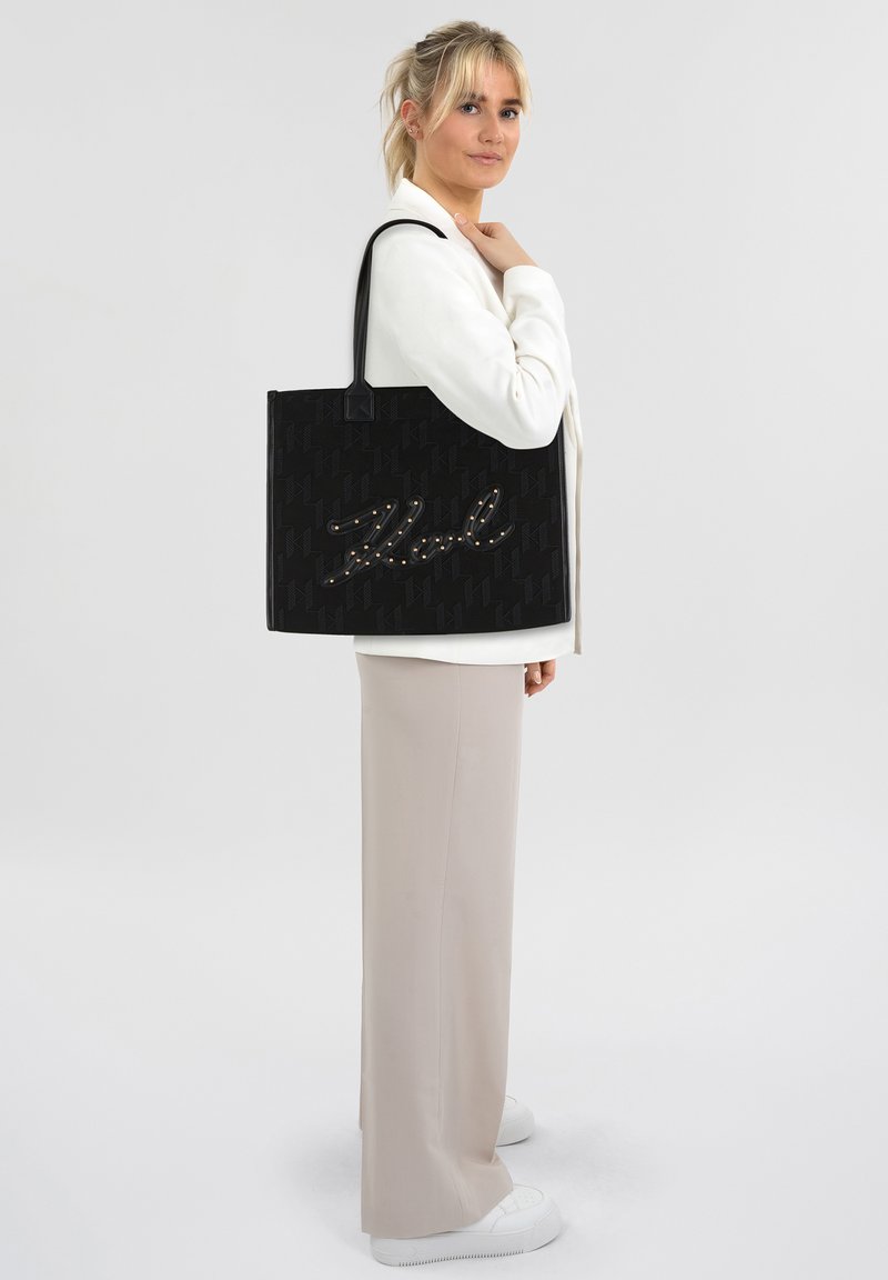 Zwarte tote bag van textuurstof met een verhoogd, schriftschriftontwerp versierd met parels. Lange zwarte banden, rechthoekige vorm.