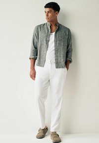 Chemise en lin grise à boutons sur un t-shirt blanc, associée à un pantalon blanc et des mocassins marron. Textures douces, coupe décontractée.