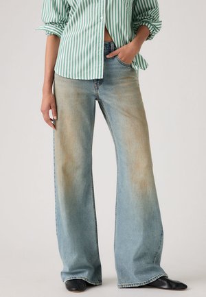 Femme portant un jean large bleu clair avec des délavages marron et des chaussures noires, associé à un chemisier rayé vert et blanc.