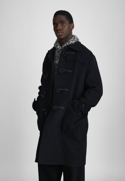 Manteau duffle noir en laine avec des fermetures à bascule, de grandes poches et un col structuré, superposé sur un pull en maille gris à motifs.