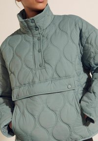 Lavet pullover i blød grøn, med høj krave, knaplukning og en forlommer med glat tekstur og rundet mønster.