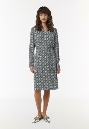 Schwarz-weiß gemustertes Kleid mit langen Ärmeln, Kragen und tailliertem Schnitt. Das Model trägt metallische flache Schuhe; glatte Stofftextur.
