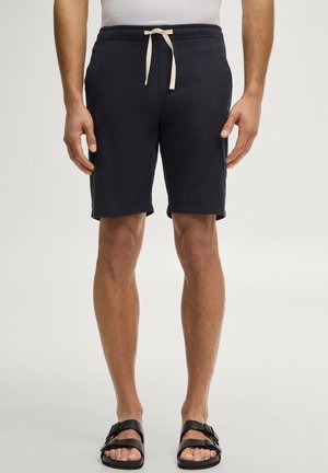 Mann trägt schwarze Shorts mit Kordelzug und schwarze Sandalen mit zwei Riemen, steht mit entspannten Armen an den Seiten vor neutralem Hintergrund.