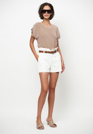 Lichtbruin gebreid bovenstuk met korte mouwen, witte shorts met hoge taille en een bruine riem. Outfit gecombineerd met beige sandalen met hak en oversized zonnebril.