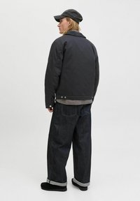 Jeune personne portant une casquette noire, une veste noire, une chemise à carreaux, un large jean foncé et des chaussures noires, se tenant de dos contre un fond uni.