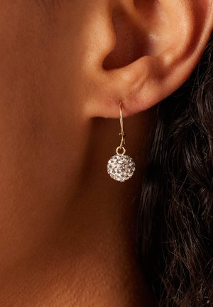 Gros plan sur une oreille portant une boucle d'oreille en or en forme de crochet avec un pendentif rond orné de cristaux qui pend.