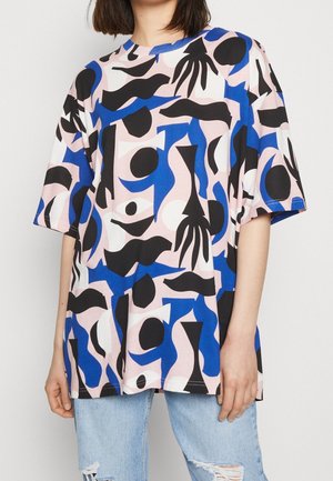 Osoba nosząca oversize T-shirt z abstrakcyjnymi czarnymi, niebieskimi, białymi i różowymi kształtami w połączeniu z jasnoniebieskimi podartymi dżinsami.