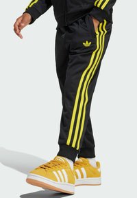 Black athletic pants with yellow-striped accents, featuring an embroidered logo. Paired with bright yellow sneakers and white soles.