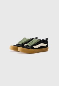 Sneakers neri in tela con lacci elastici verdi, striscia bianca laterale e suola in gomma naturale. Presentano una punta rinforzata e materiali tessili.