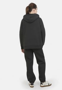 Sudadera negra de gran tamaño con capucha ajustable, combinada con pantalones deportivos a juego. Tejido suave con puños acanalados; calzado casual con detalles en blanco y negro.