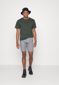 Donkergroene t-shirt met korte mouwen, grijze geruite shorts en zwarte veterschoenen. De look wordt compleet gemaakt met een zwarte bucket hat.