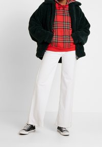 Giacca in pile verde, felpa con motivo a quadri rossi, jeans bianchi a gamba larga, sneaker nere. L'outfit presenta una giacca testurizzata e una felpa con un motivo.