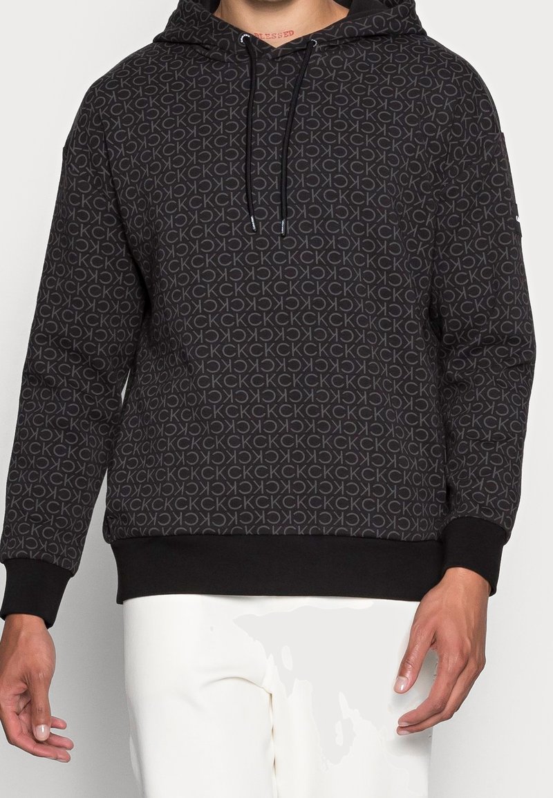 Sudadera negra con un patrón repetido de logotipos "CK" en gris claro, capucha con cordón y puños y dobladillo acanalados. Textura de tela suave.