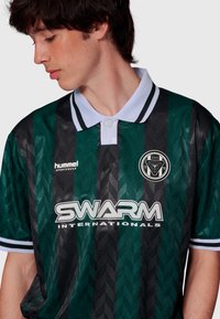 Kortärmad grön- och svartrandig pikétröja med ett texturerat mönster, vit krage och SWARM-logotyp. Tillverkad av lätt tyg.