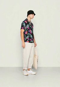 Cămașă neagră cu mânecă scurtă, având un model floral colorat, asociată cu pantaloni bej deschis și adidași albi, purtând o geantă de umăr bej.