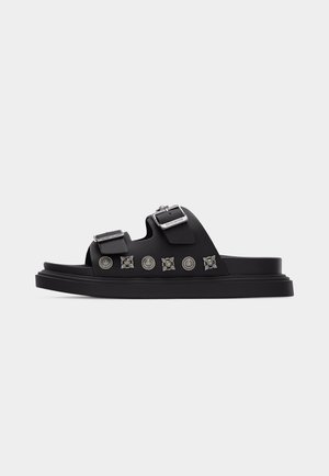 Μαύρο σανδάλι με πλάγια σφήνα, δύο ρυθμιζόμενες αγκράφες και διακοσμητικά ασημένια καρφιά στο πλάι, με επίπεδη σόλα.