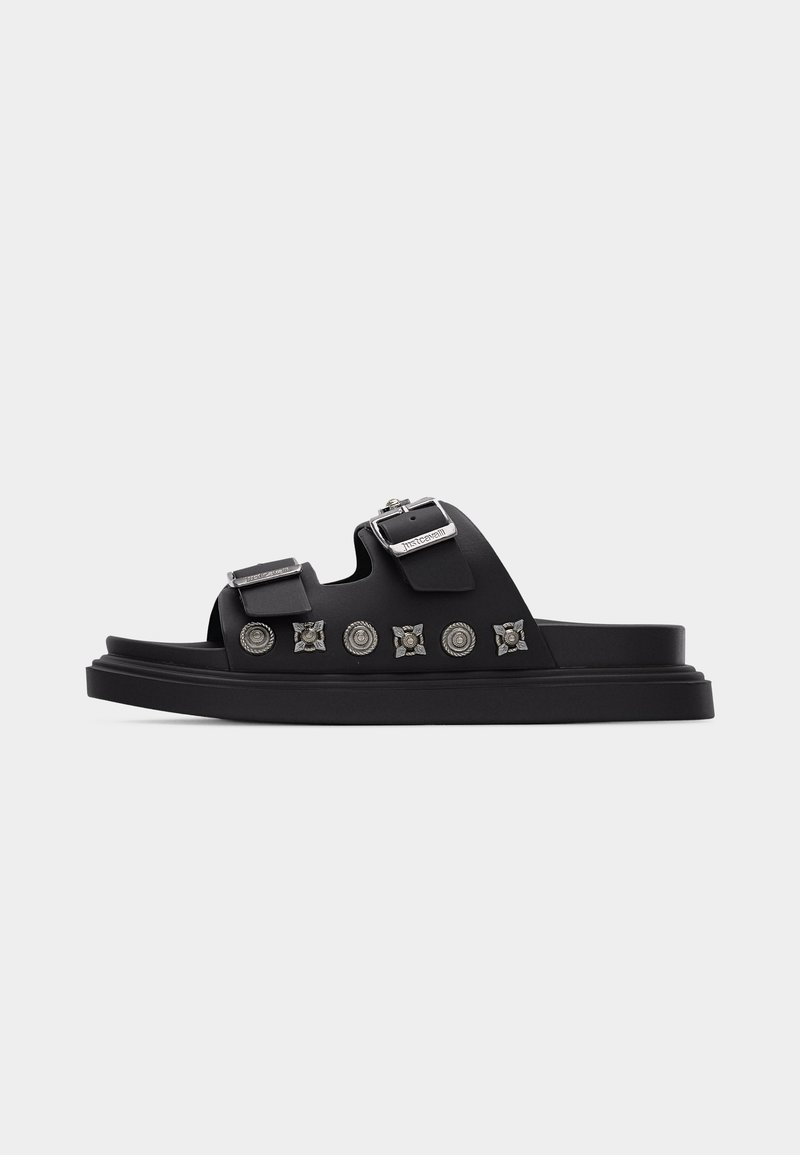 Μαύρο σανδάλι με πλάγια σφήνα, δύο ρυθμιζόμενες αγκράφες και διακοσμητικά ασημένια καρφιά στο πλάι, με επίπεδη σόλα.