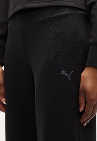 Μαύροι υφασμάτινοι παντελόνες με μικρό λογότυπο Puma στο αριστερό μηρό, που φοριούνται από ένα άτομο με το δεξί του χέρι χαλαρά στο πλάι.