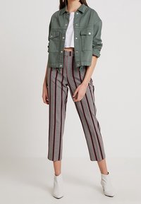 Kvinna i grön kort jacka, vit kort topp, mönstrade högmidjade byxor med ränder och vita ankelstövlar, stående mot en enhetlig bakgrund.