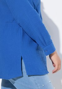Camicia blu, con texture e polsini con bottoni e spacchi laterali, realizzata in un tessuto leggero, abbinata a jeans azzurri arricchiti da perle.