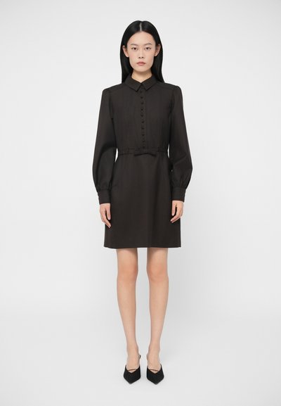 Femme se tenant de face, portant une robe noire à manches longues avec des boutons à l'avant, un col, un nœud à la taille et des escarpins noirs pointus.