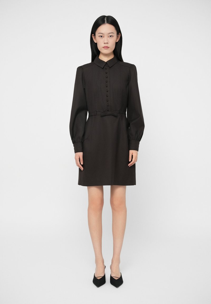 Sieviete stāv profila pozīcijā, uzvilkusi melnu garām piedurknēm tērpu ar priekšējiem pogām, apkakli, jostu vidukļa daļā un melnām smailajām zābaku kurpēm.