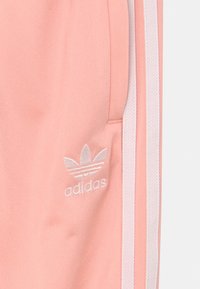 Světle růžové sportovní kalhoty vyrobené z hladkého materiálu, s bílými bočními pruhy a vyšitým logem na přední levé straně.