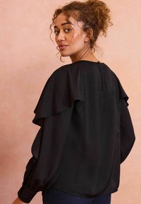 Blusa nera con design a strati e volant sulle maniche, scollatura tonda con chiusura a bottone sul retro e texture liscia.