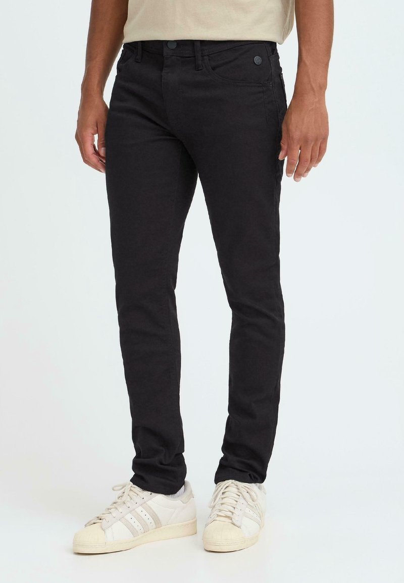 Homme portant un pantalon noir slim-fit et des baskets blanches, debout devant un fond clair uni.