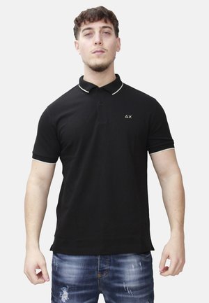 Giovane uomo in piedi, indossa una polo nera a maniche corte con rifiniture bianche e jeans blu consumati su uno sfondo semplice.