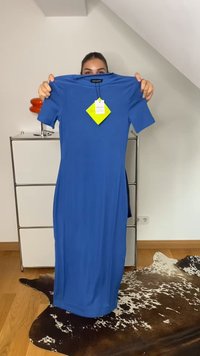 Eine Person hält ein blaues Kurzarmkleid mit einem gelben Etikett in einem Raum mit einem Kuhhautteppich und einem weißen Aufbewahrungsschrank hoch.